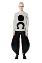 A person standing wearing a white long-sleeve jumper with a geometric logo on the front and black trousers.