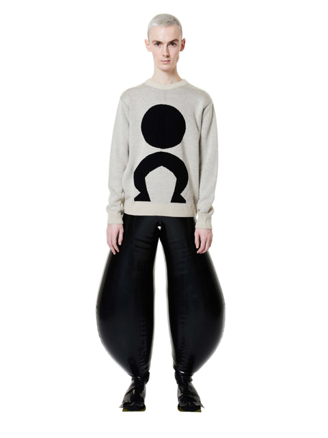 A person standing wearing a white long-sleeve jumper with a geometric logo on the front and black trousers.