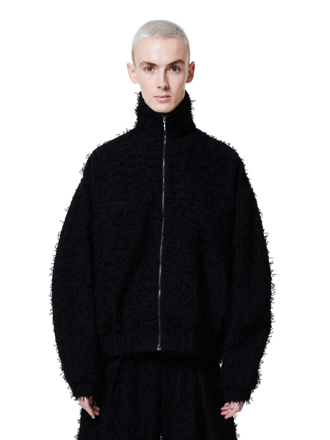 A male model wearing a black, fuzzy textured jacket with a standing collar and a front zip closure.