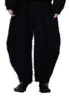 A person standing wearing wide-leg black trousers with a fuzzy texture, designed to be worn loose, with the trousers tapering at the ankle.